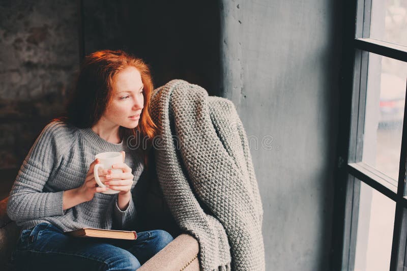 Glückliche Rothaarigefrau, die sich zu Hause im gemütlichen Winter- oder Herbstwochenende mit dem Buch und Schale heißem Tee, sit