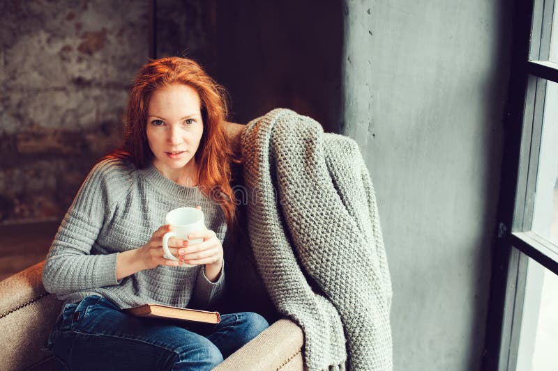 Glückliche Rothaarigefrau, die sich zu Hause im gemütlichen Winter- oder Herbstwochenende mit dem Buch und Schale heißem Tee, sit stockfotografie