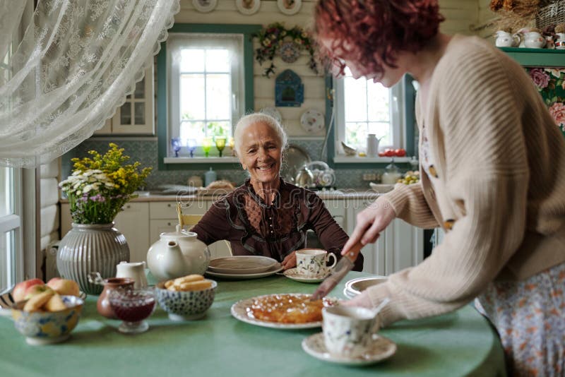 Glückliche Seniorin sitzt am Tisch und schaut Mädchen schneidend Kuchen lizenzfreies stockbild