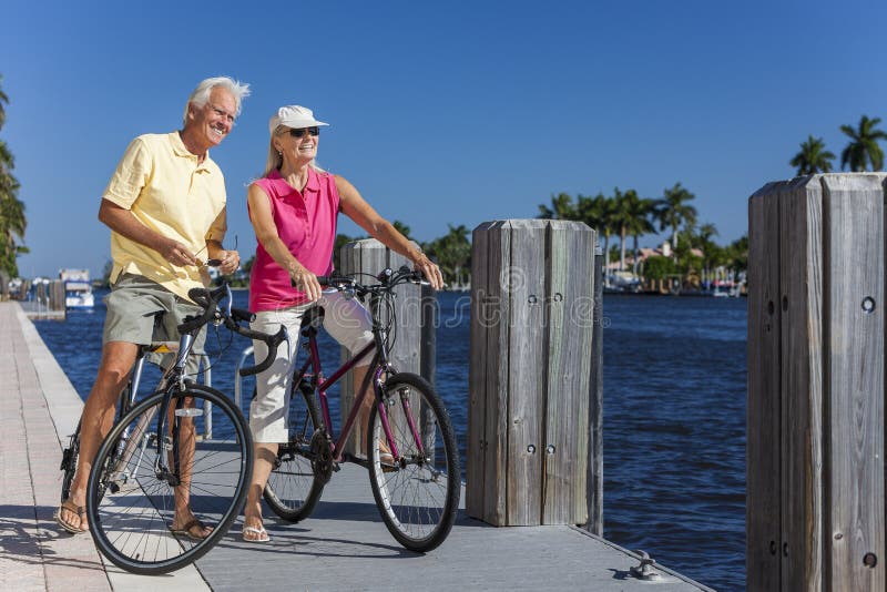 Happy senior men and women couple together cycling on bicycles with bright clear blue sky by a river or sea. Happy senior men and women couple together cycling on bicycles with bright clear blue sky by a river or sea