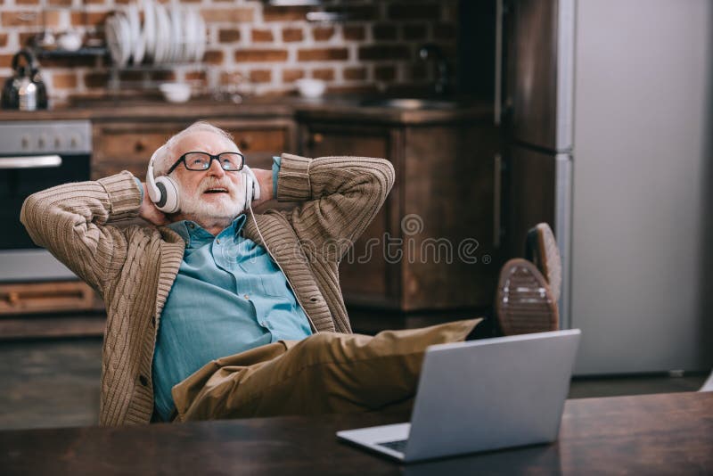 Glücklicher alter Mann in den Kopfhörern unter Verwendung des Laptops mit Füßen lizenzfreies stockfoto Glücklicher alter Mann in den Kopfhörern unter Verwendung des Laptops mit Füßen lizenzfreies stockfoto