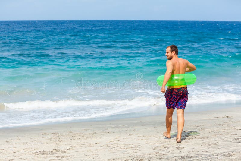 Glücklicher Barthipster in aufblasbarem Ring macht Spaß beim Strandspaziergang. Der Sonnenmann läuft am Meeresufer entlang und spr lizenzfreie stockfotografie