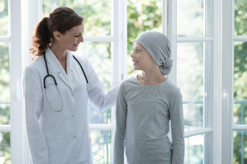 Glücklicher Doktor, der positives Kind mit tragendem Kopftuch Krebses stützt lizenzfreie stockfotografie Glücklicher Doktor, der positives Kind mit tragendem Kopftuch Krebses stützt lizenzfreie stockfotografie
