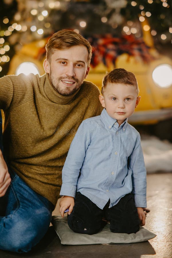 Glücklicher Vater mit seinem Sohn sitzen auf dem Boden gegen Hintergrund der glühenden Scheinwerfern und Weihnachtsdekorationen lizenzfreies stockbild