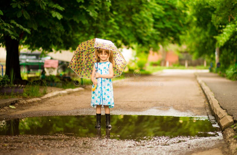Glückliches Kindermädchen im Kleid mit einem Regenschirm und Gummistiefeln in der Pfütze auf Weg lizenzfreie stockbilder Glückliches Kindermädchen im Kleid mit einem Regenschirm und Gummistiefeln in der Pfütze auf Weg lizenzfreie stockbilder