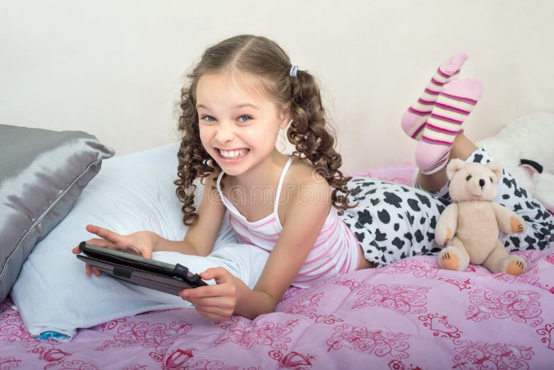 Glückliches kleines Mädchen, das auf Bett mit Tablet-Computer liegt stockfotos