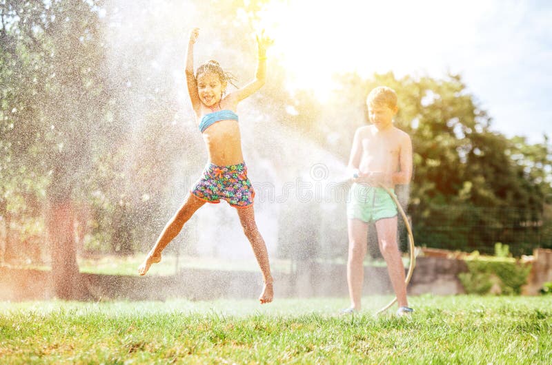 Glückliches kleines Mädchen springt unter Wasser, wenn Bruder sie vom Gartenschlauch gießt Heiße Sommertagestätigkeit