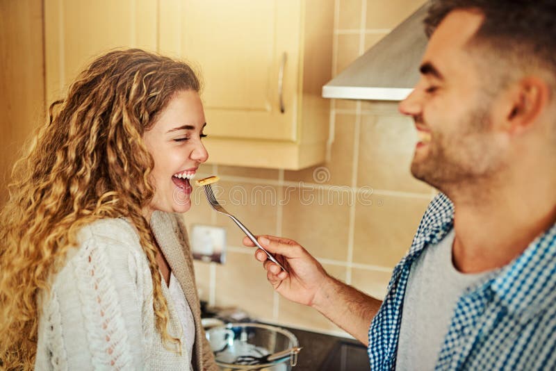 Glückliches Paar lacht und füttert mit Frühstück in der Küche für morgendliche Biss Bindung oder Geschmack zu Hause. junger Mann u stockfotos