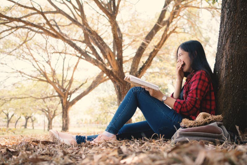 Godere della donna dei pantaloni a vita bassa di momento che legge un libro e che si siede sotto il grande albero sul parco fotografia stock Godere della donna dei pantaloni a vita bassa di momento che legge un libro e che si siede sotto il grande albero sul parco fotografia stock