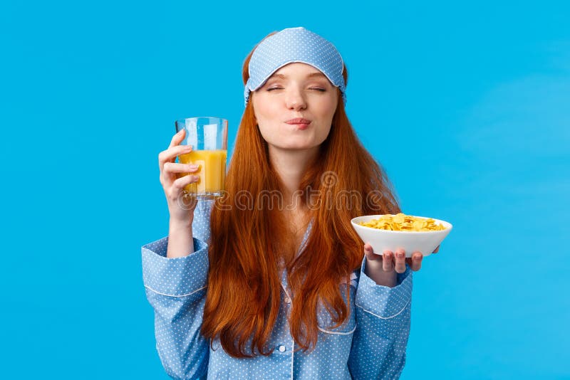 Morning, healthy lifestyle and people concept. Feminine good-looking redhead woman in sleep mask and pyjama, close eyes and licking lips from satisfaction, eating breakfast, orange juice, cereals. Morning, healthy lifestyle and people concept. Feminine good-looking redhead woman in sleep mask and pyjama, close eyes and licking lips from satisfaction, eating breakfast, orange juice, cereals.