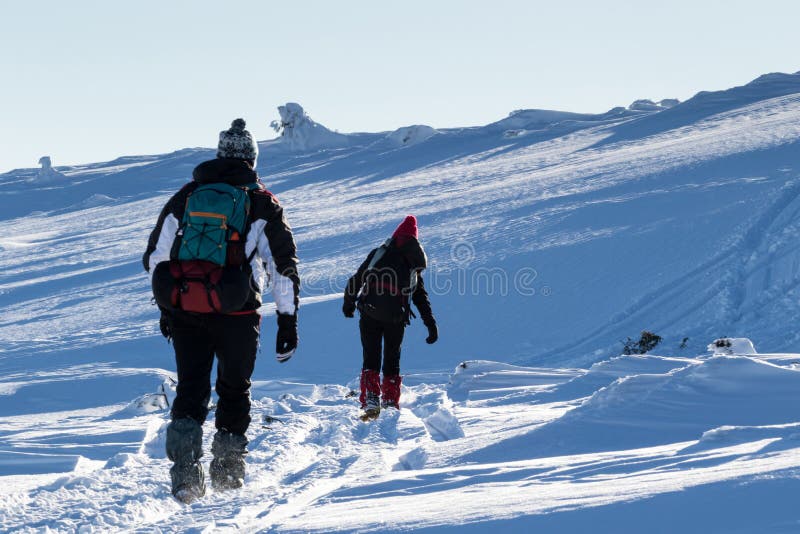 A young couple are trekking through frozen snow on top of a mountain during a sunny day of winter