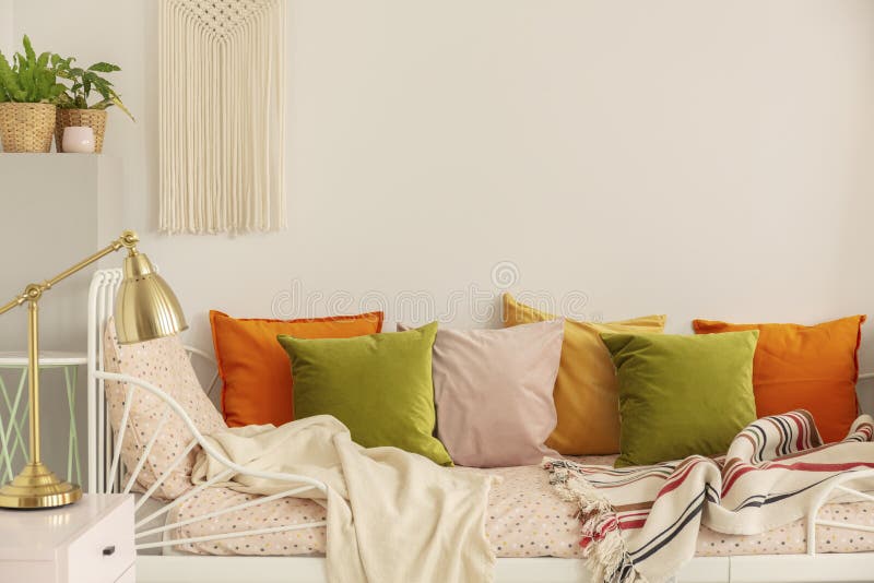 Golden lamp on the nightstand next to olive green, pastel pink, yellow and orange pillows on single metal bed with patterned