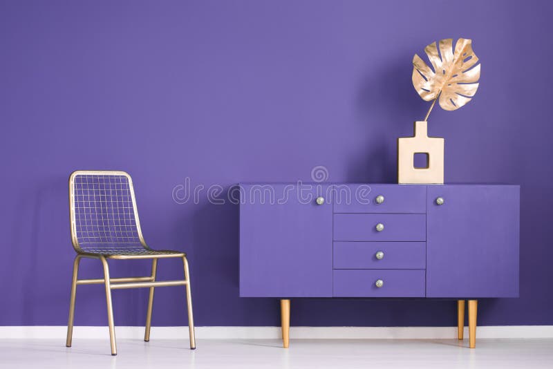 Golden monstera leaf in a vase standing on an ultra violet cupboard next to a metal chair in creative living room interior. Real