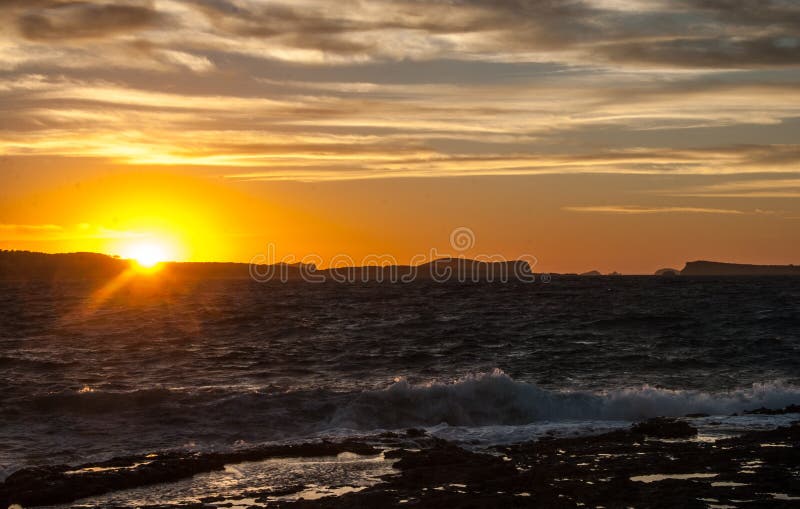 Golden sunset behind Conejera Islands. Choppy waters of Balearic sea churns waves on rocks along shore. View of Conejera Islands from behind cafe del mar in Ibiza in November.