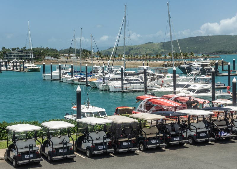 Hamilton Island, Australia - February 16, 2019: Row of golf carts as regular street vehicles parked at yacht harbor on marina. Azure water, white boats, green hills, blue sky. Hamilton Island, Australia - February 16, 2019: Row of golf carts as regular street vehicles parked at yacht harbor on marina. Azure water, white boats, green hills, blue sky