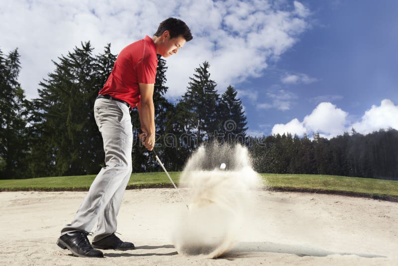 Young male golf player in red shirt and grey pants chipping golf ball out of a sand trap with sand wedge and sand caught in motion. Young male golf player in red shirt and grey pants chipping golf ball out of a sand trap with sand wedge and sand caught in motion.