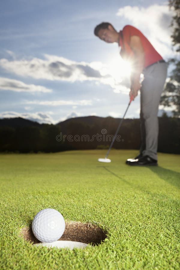 Smiling professional male golf player putting successfully ball on green with close up of ball dropping into cup. Smiling professional male golf player putting successfully ball on green with close up of ball dropping into cup.