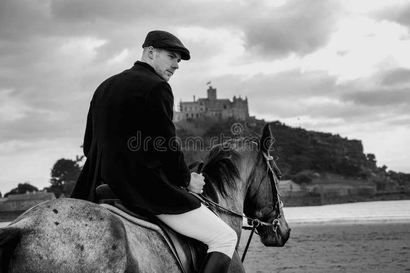 Good Looking Male Horse Rider riding horse on beach in traditional riding clothing with St Michael`s Mount in background
