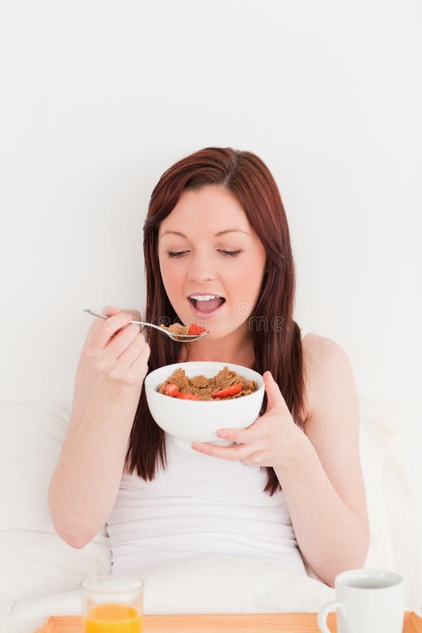 Good looking red-haired female having her breakfast while sitting on her bed