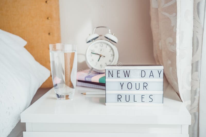 Good morning concept. Inspiration Motivational Life Quotes New day your rules message on lightened box with alarm clock, notebooks and glass of water on the bedside table in the sunlight. Copy space