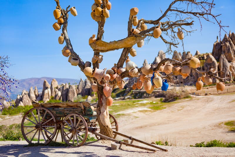 Goreme National Park, Cappadocia, Turkey. Old wooden cart stands under dead tree with clay pots hanging from its branches. Unusual landscape with conical rocks is in blurred background. Goreme National Park, Cappadocia, Turkey. Old wooden cart stands under dead tree with clay pots hanging from its branches. Unusual landscape with conical rocks is in blurred background