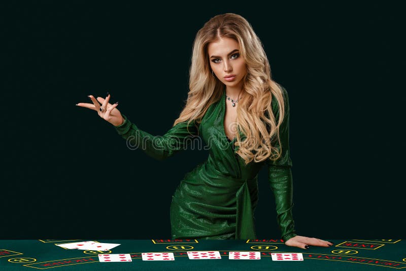 Girl in stylish dress is showing something, leaning on playing table with cards on it, posing on green background. Poker