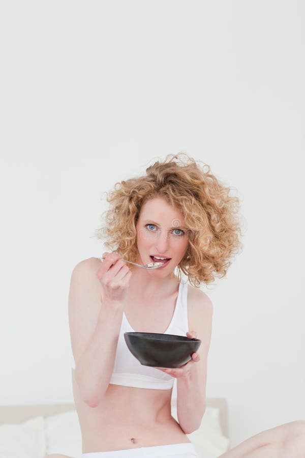 Gorgeous blonde woman eating a bowl of cereals while sitting on her bed in her appartment