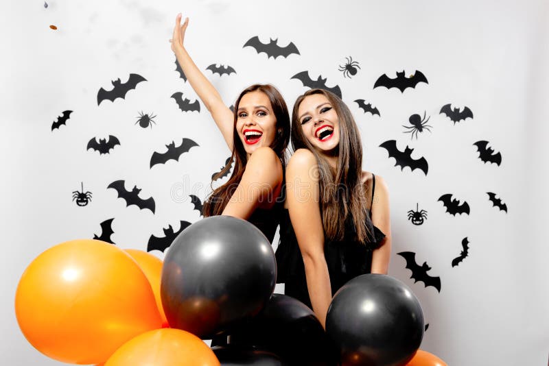 Gorgeous brunette women in black dress holds black and orange balloons on a white background with black bats and spiders. Halloween .