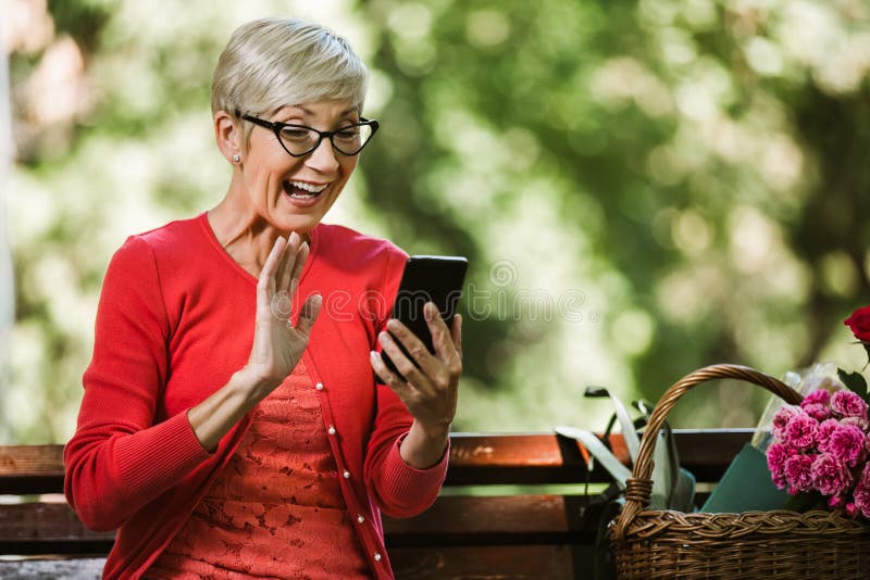 Caucasian senior woman with blonde short hair sitting on bench in park and make video call with smart phone