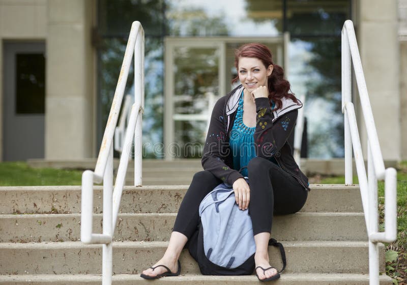 Beautiful female student, with red hair, wearing backpack on campus - sitting on stairs. Beautiful female student, with red hair, wearing backpack on campus - sitting on stairs