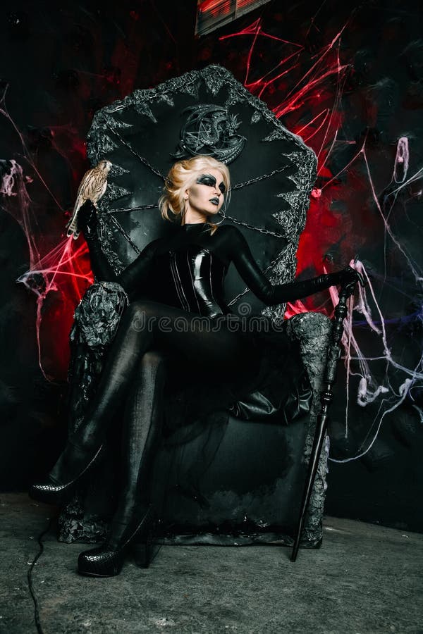 Gothic girl with scary makeup on a huge throne ready for Halloween party. Young blonde woman with little bird on a black chair