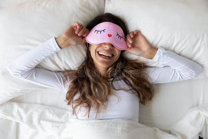 Above top view head shot happy millennial mixed race healthy woman lying in bed with sleeping mask, waking up after sweet dreams. Smiling young relaxed lady feeling energetic in morning at home. Above top view head shot happy millennial mixed race healthy woman lying in bed with sleeping mask, waking up after sweet dreams. Smiling young relaxed lady feeling energetic in morning at home.