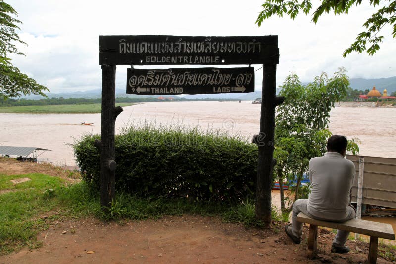 Chiang Saen, Thailand - August 04 2012: Old Thai sat next to a sign at the Golden Triangle indicating the directions of the Thai and Laos borders. On the opposite bank of the Mekong river, is the Golden Triangle Special Economic Zone in Laos. This special economic zone is a 99-year lease made from the Laotian government to the Chinese-owned Kings Romans Group. Chiang Saen, Thailand - August 04 2012: Old Thai sat next to a sign at the Golden Triangle indicating the directions of the Thai and Laos borders. On the opposite bank of the Mekong river, is the Golden Triangle Special Economic Zone in Laos. This special economic zone is a 99-year lease made from the Laotian government to the Chinese-owned Kings Romans Group.