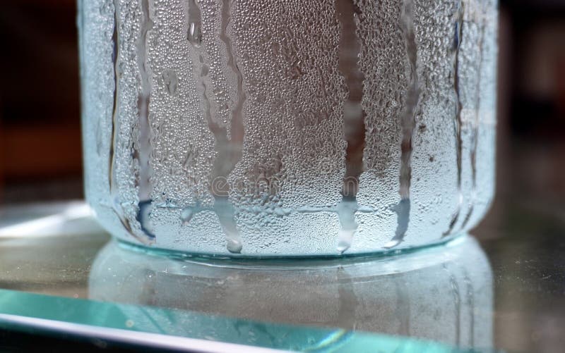 Cold temperature water in clear water bottle with humid moisture water drips on the outside of the container isolated on horizontal glass table background. Cold temperature water in clear water bottle with humid moisture water drips on the outside of the container isolated on horizontal glass table background.