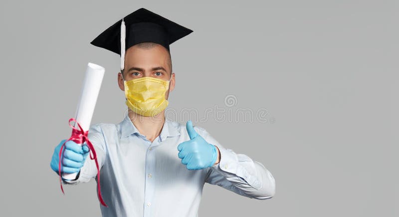 High school graduation during a quarantine. Student graduate in a hat and a protective mask holds a diploma and shows thumb up. High school graduation during a quarantine. Student graduate in a hat and a protective mask holds a diploma and shows thumb up