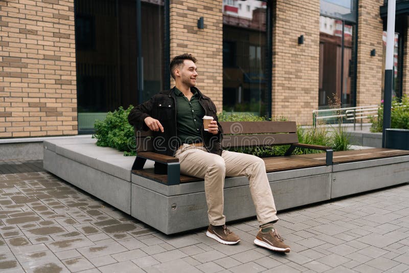 Grand cliché d'un homme heureux et détendu assis sur le banc de la ville à l'extérieur par un bâtiment en briques savourant une ta photo libre de droits