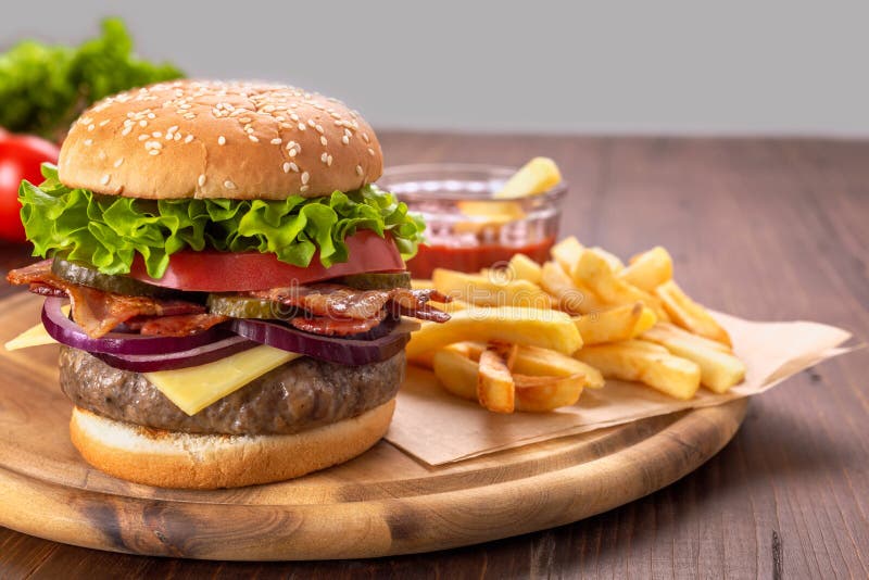 Grand hamburger délicieux avec les fritures et le ketchup. Hamburger avec Fried Bacon, les conserves au vinaigre coupées en tranches, le fromage, l'oignon rouge, les tomates et la laitue sur un plan rapproché de planche à découper avec les pommes frites et le ketchup sur un Tableau en bois