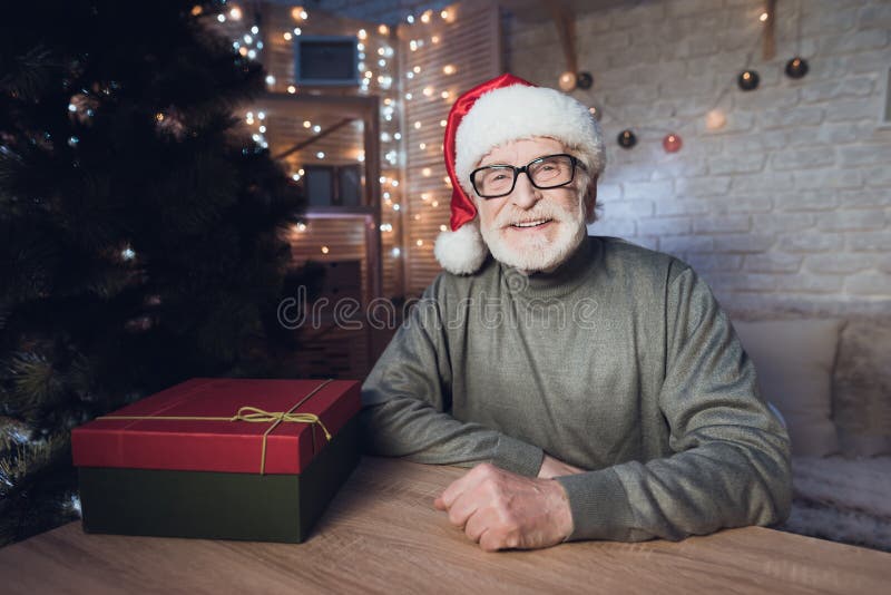 Grandfather is sitting near Cristmas tree in Santa Claus`s hat at night at home.