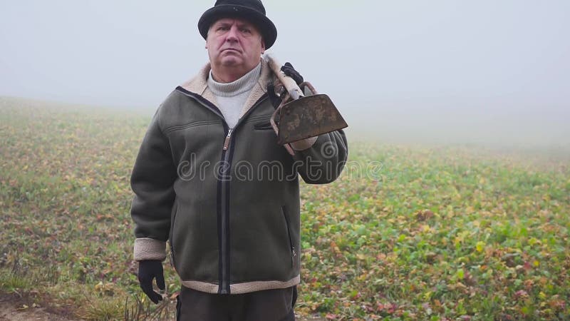 Granjero mayor con el sombrero que sostiene una azada y un bieldo fangosos en el campo Trabajo manual niebla