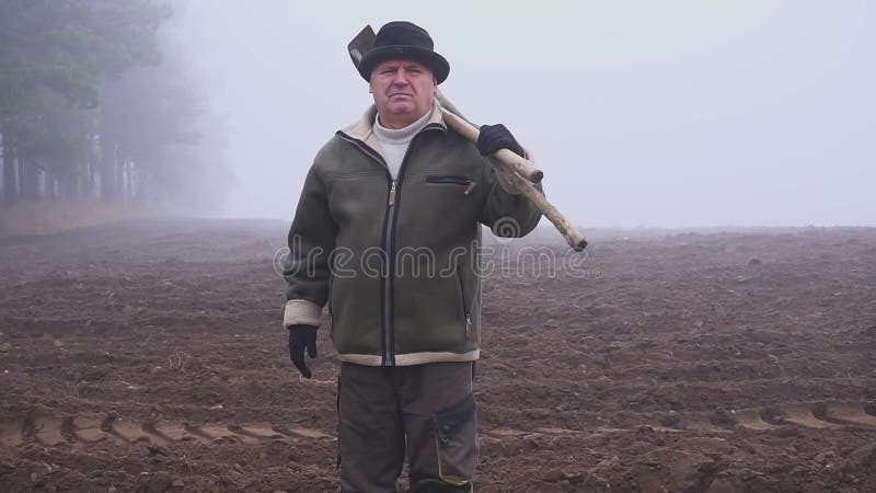 Granjero mayor con el sombrero que sostiene una azada y un bieldo fangosos en el campo Trabajo manual niebla