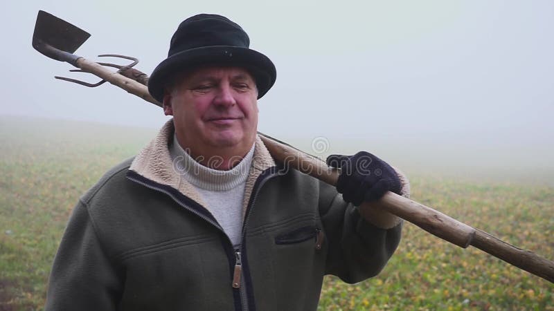 Granjero mayor con el sombrero que sostiene una azada y un bieldo fangosos en el campo Trabajo manual niebla