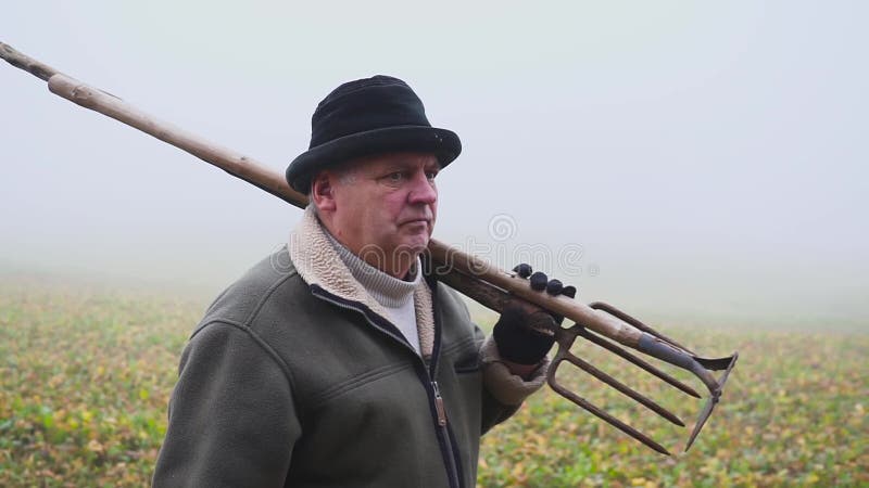 Granjero mayor con el sombrero que sostiene una azada y un bieldo fangosos en el campo Trabajo manual niebla