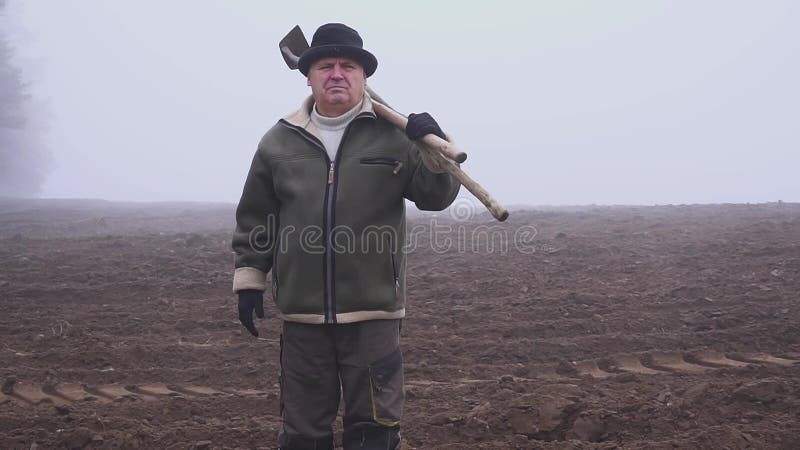 Granjero mayor con el sombrero que sostiene una azada y un bieldo fangosos en el campo Trabajo manual niebla