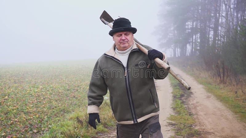 Granjero mayor con el sombrero que sostiene una azada y un bieldo fangosos en el campo Trabajo manual niebla