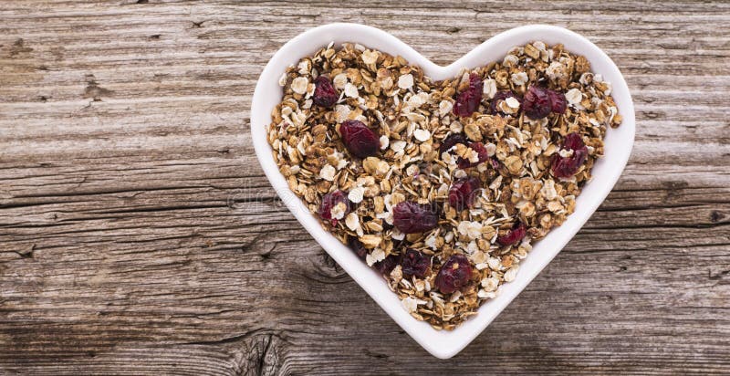 Home caramel granola with dried cranberries in a white ceramic dish in the shape of heart on the texture wooden background. Top view. Horizontal. The concept of organic food. selective Focus. Home caramel granola with dried cranberries in a white ceramic dish in the shape of heart on the texture wooden background. Top view. Horizontal. The concept of organic food. selective Focus