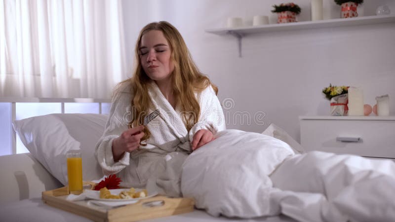 Grappig meisje die licht ontbijt eten, die in bed in hotelruimte zitten, die honger voelen royalty-vrije stock foto's Grappig meisje die licht ontbijt eten, die in bed in hotelruimte zitten, die honger voelen royalty-vrije stock foto's