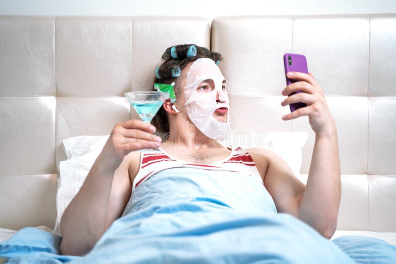 Funny man with a cosmetic mask on his face and curlers does spa treatments in a room holding cocktail in his hand spends a stream for social networks. Parody on cosmetics beauty and fashion bloggers. Funny man with a cosmetic mask on his face and curlers does spa treatments in a room holding cocktail in his hand spends a stream for social networks. Parody on cosmetics beauty and fashion bloggers