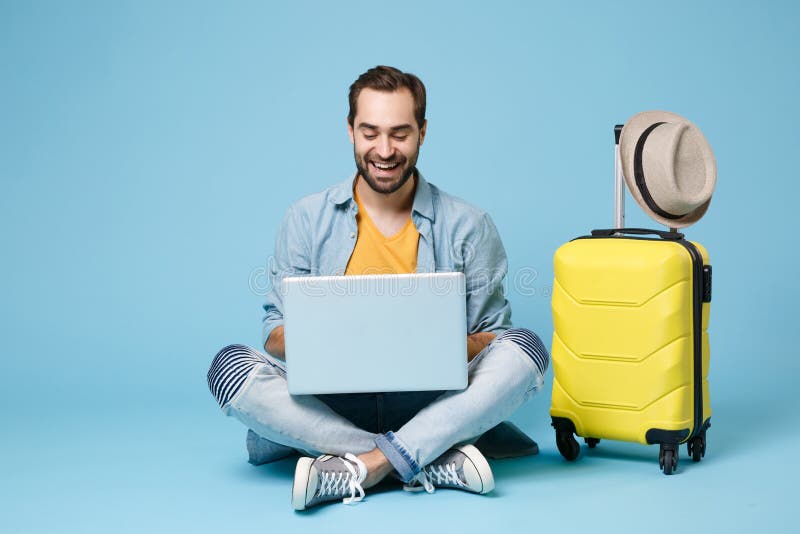 Funny traveler tourist man in yellow casual clothes isolated on blue background. Passenger traveling abroad on weekend. Air flight journey concept. Sit near suitcase, work on laptop, booking hotel. Funny traveler tourist man in yellow casual clothes isolated on blue background. Passenger traveling abroad on weekend. Air flight journey concept. Sit near suitcase, work on laptop, booking hotel