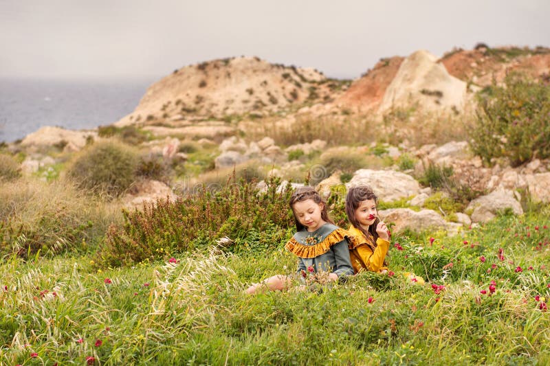 On the grass plateau sit two sisters girls in retro clothes are considered sniffing flowers with hills and stormy sea on a backgro royalty free stock photography On the grass plateau sit two sisters girls in retro clothes are considered sniffing flowers with hills and stormy sea on a backgro royalty free stock photography
