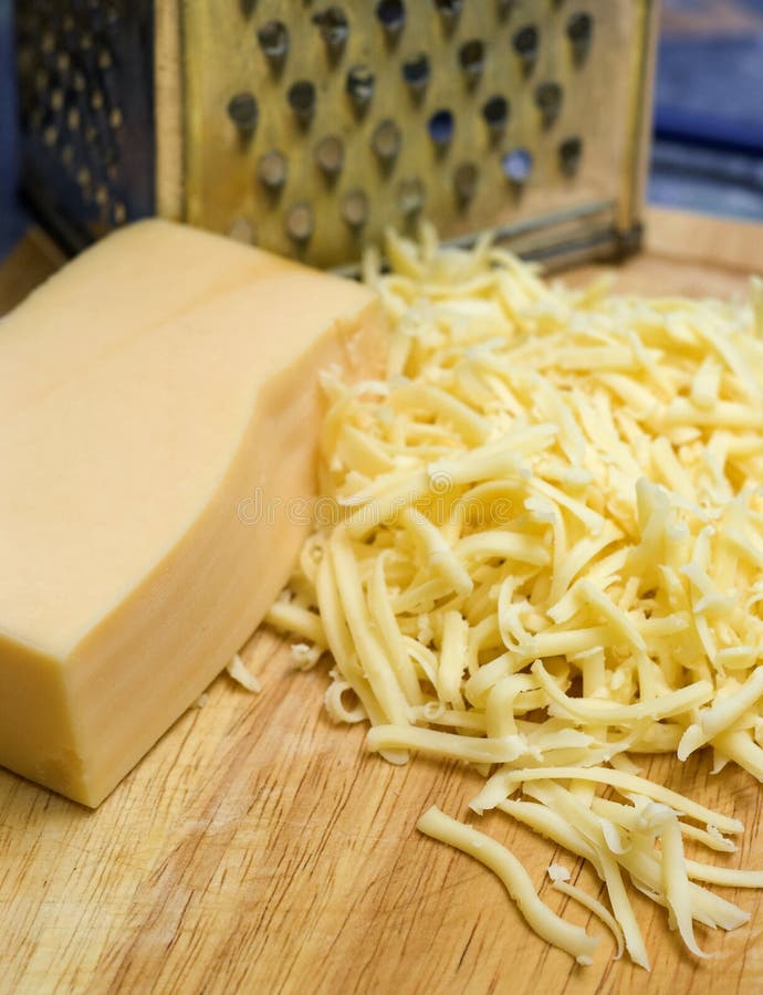 Grating cheese. A hunk of yello cheese and shredded pieces, metal grater in the background Grating cheese. A hunk of yello cheese and shredded pieces, metal grater in the background
